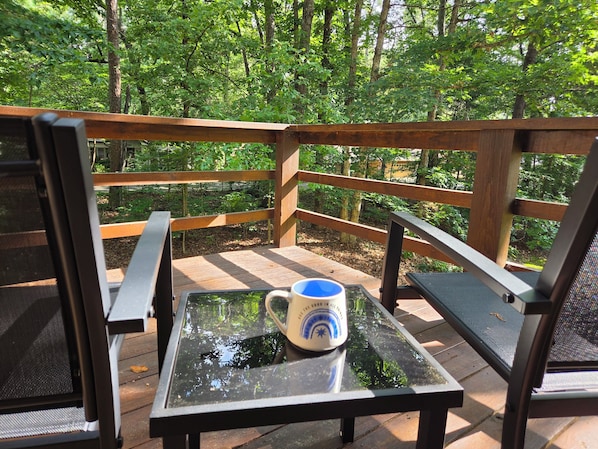 A wooden deck with a glass table and chairs, surrounded by a forest.