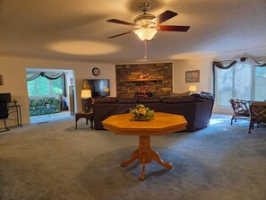 A living room with a stone fireplace, a ceiling fan, a television, and a dining table with a vase of flowers. - West Gate Retreat 3 bedroom home near Desoto Golf Club in Hot Springs Village (Hot Springs)