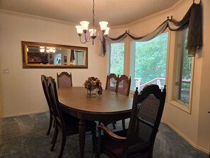 A dining room with a round table, six chairs, a chandelier, a mirror, and a window with curtains.
