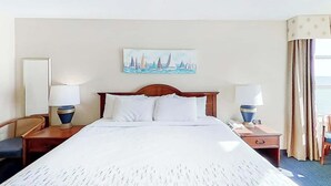 A hotel room with a large bed, two bedside tables with lamps, a chair, a painting on the wall, and a window with curtains. - Carolinian Beach Resort 0830 (Myrtle Beach)