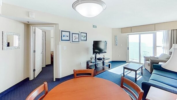 A hotel room with a dining table, chairs, a TV, a sofa, and a large window with a view. - Carolinian Beach Resort 0830 (Myrtle Beach)