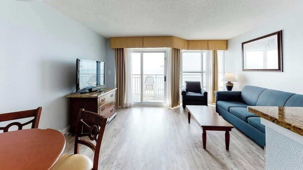Ein Hotelzimmer mit Esstisch, einer Couch, einem Fernseher und einem Balkon mit Vorhängen.
