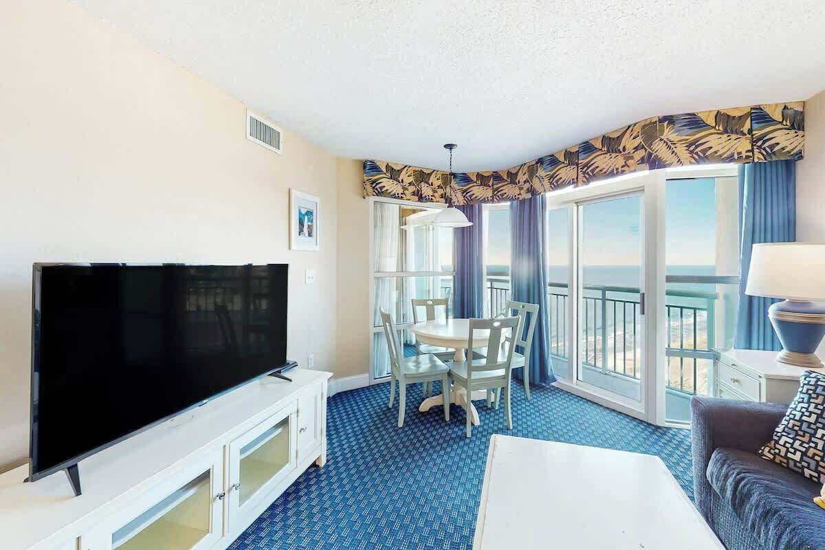 A hotel room with a flat-screen TV, a dining table with chairs, a balcony with a view, and a sofa.