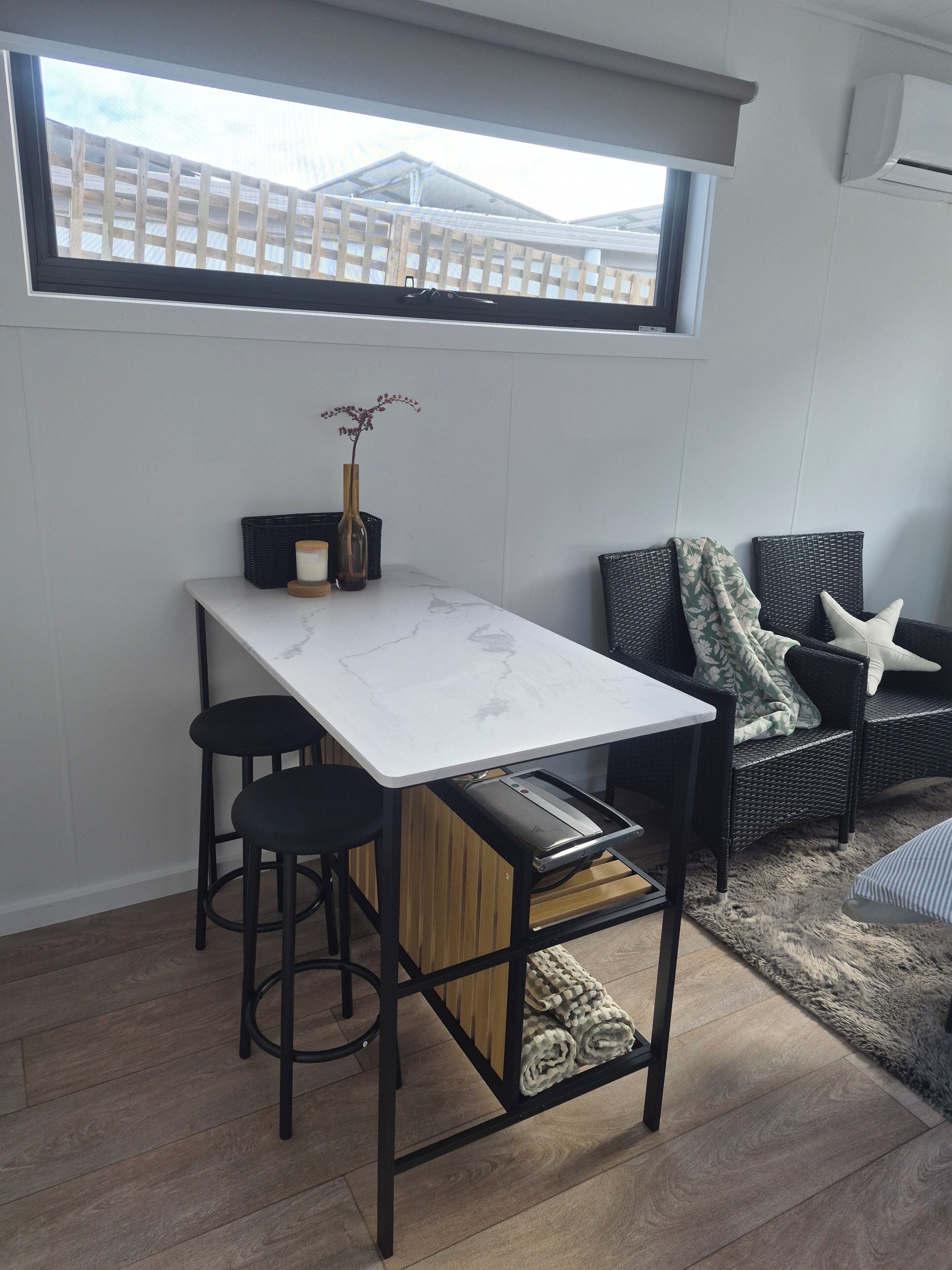 A compact room with a dining table, two stools, a sofa, and a window overlooking a wooden deck.