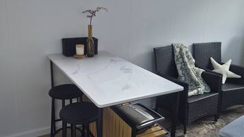 A compact room with a dining table, two stools, a sofa, and a window overlooking a wooden deck.