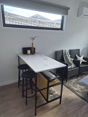 A compact room with a dining table, two stools, a sofa, and a window overlooking a wooden deck.