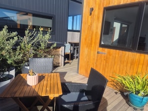 An outdoor patio area with a wooden table and chairs, potted plants, and a view of the building's exterior.