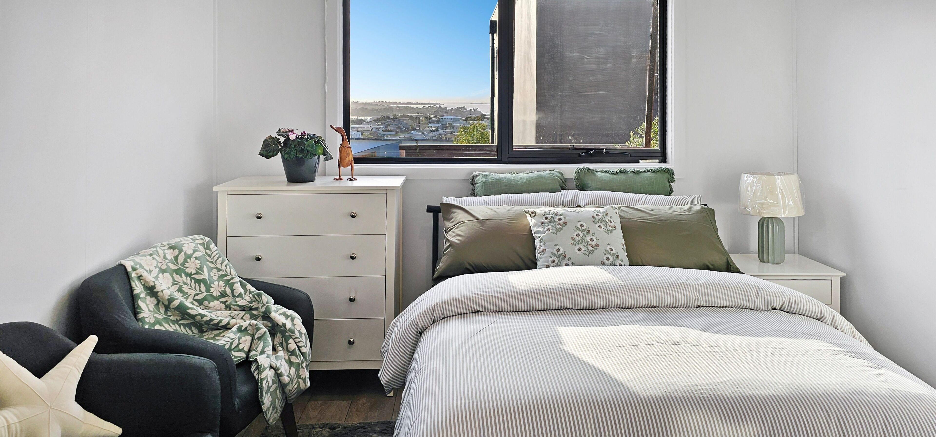 A bedroom with a bed, a chair, a dresser, a window with a view, and a lamp on a bedside table.