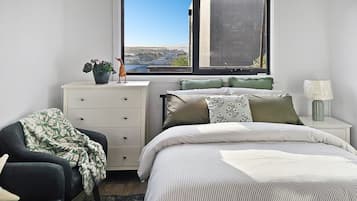 A bedroom with a bed, a chair, a dresser, a window with a view, and a lamp on a bedside table.