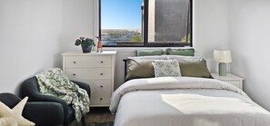 Une chambre à coucher comprenant un lit, une chaise, une commode, une fenêtre donnant sur un paysage agréable et une lampe sur une table de chevet.