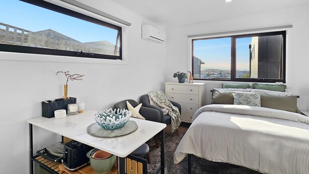 A modern bedroom with a bed, a desk, a chair, and a dresser. There are two windows providing views of the outside.
