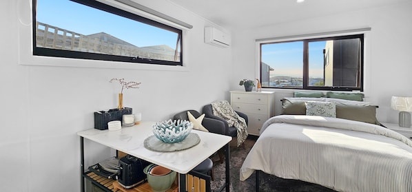A modern bedroom with a bed, a desk, a chair, and a dresser. There are two windows providing views of the outside.