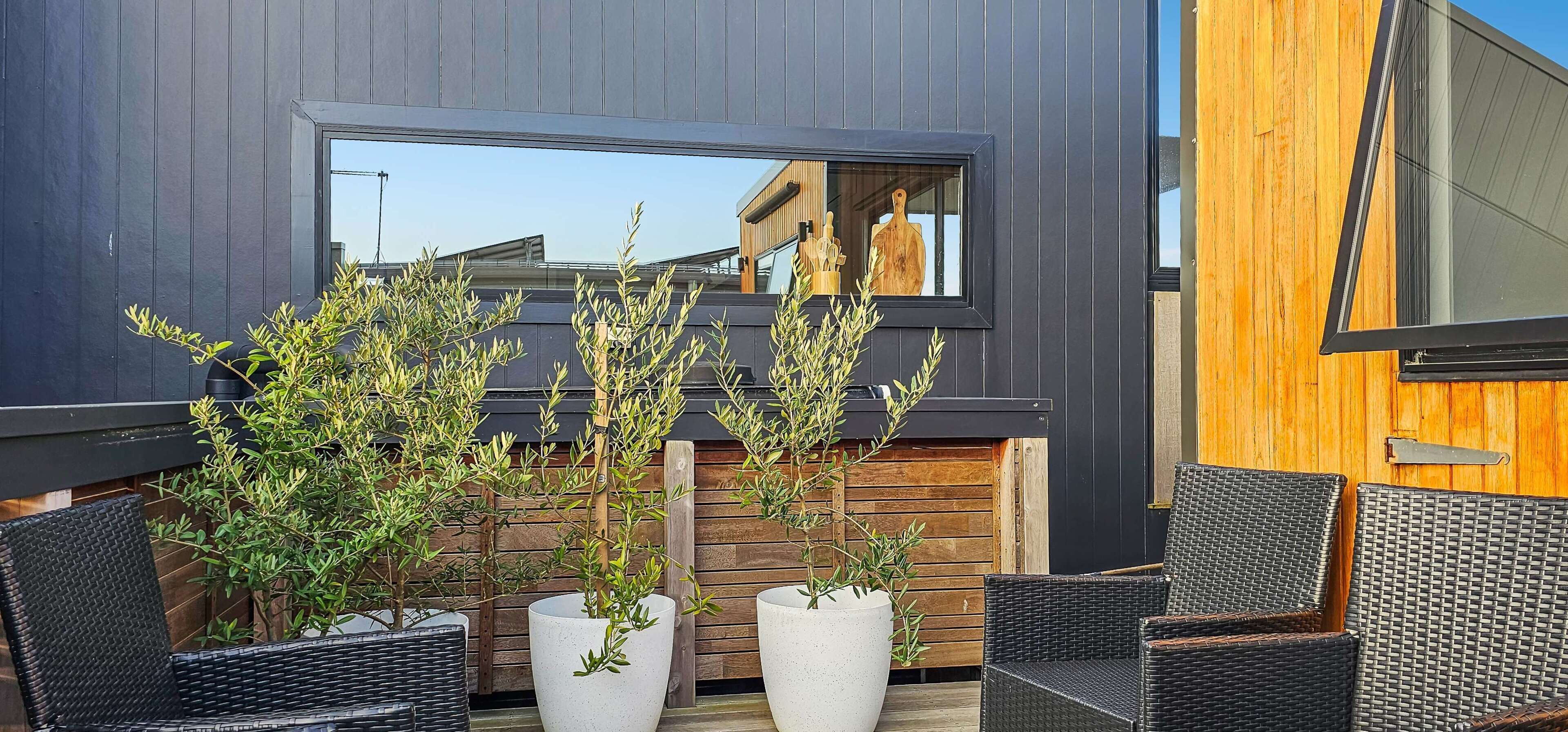 Un espace extérieur aménagé avec des chaises en osier et des plantes en pot, un mur en bois et une fenêtre qui reflète le ciel.