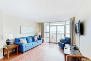 A hotel room with a blue sofa, a desk with a laptop, a TV on the wall, and a balcony with a view. - Carolinian Beach Resort 0832 (Myrtle Beach)