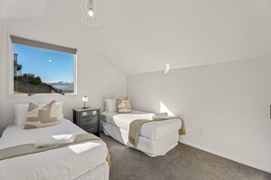 A bedroom with two beds, a nightstand, a lamp, and a view of a mountainous landscape through a window. - Panorama Point - Amazing Views & Location (Queenstown)