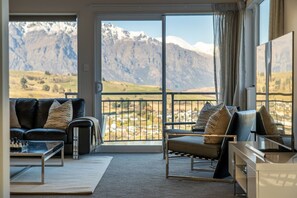 Ein Wohnzimmer mit Sofa, Sessel und Couchtisch, ein großes Fenster mit Blick auf die Berge und ein Balkon mit Geländer.