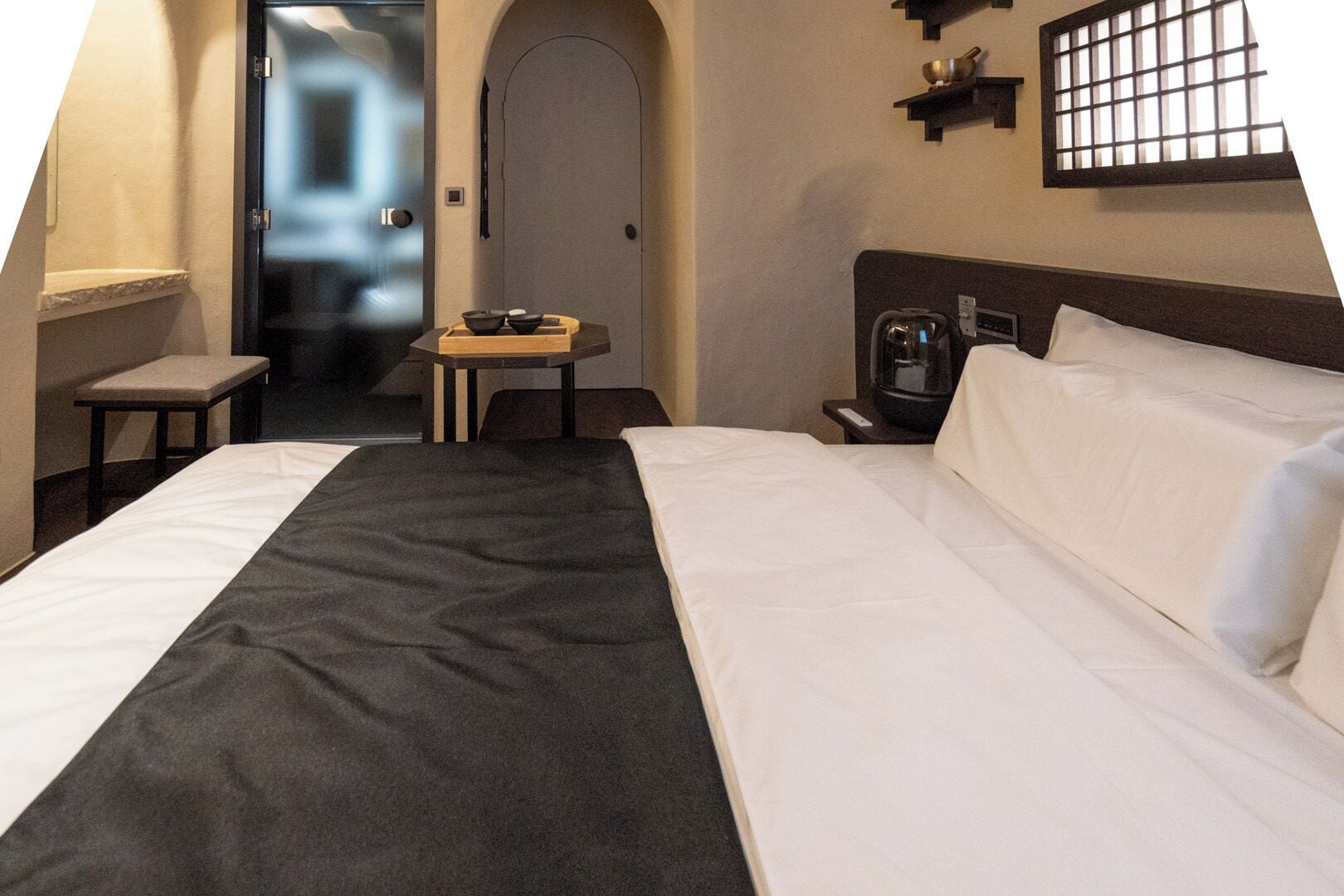 A hotel room with a bed, a small table with a bowl and a book, a coffee machine, and a window with bars.