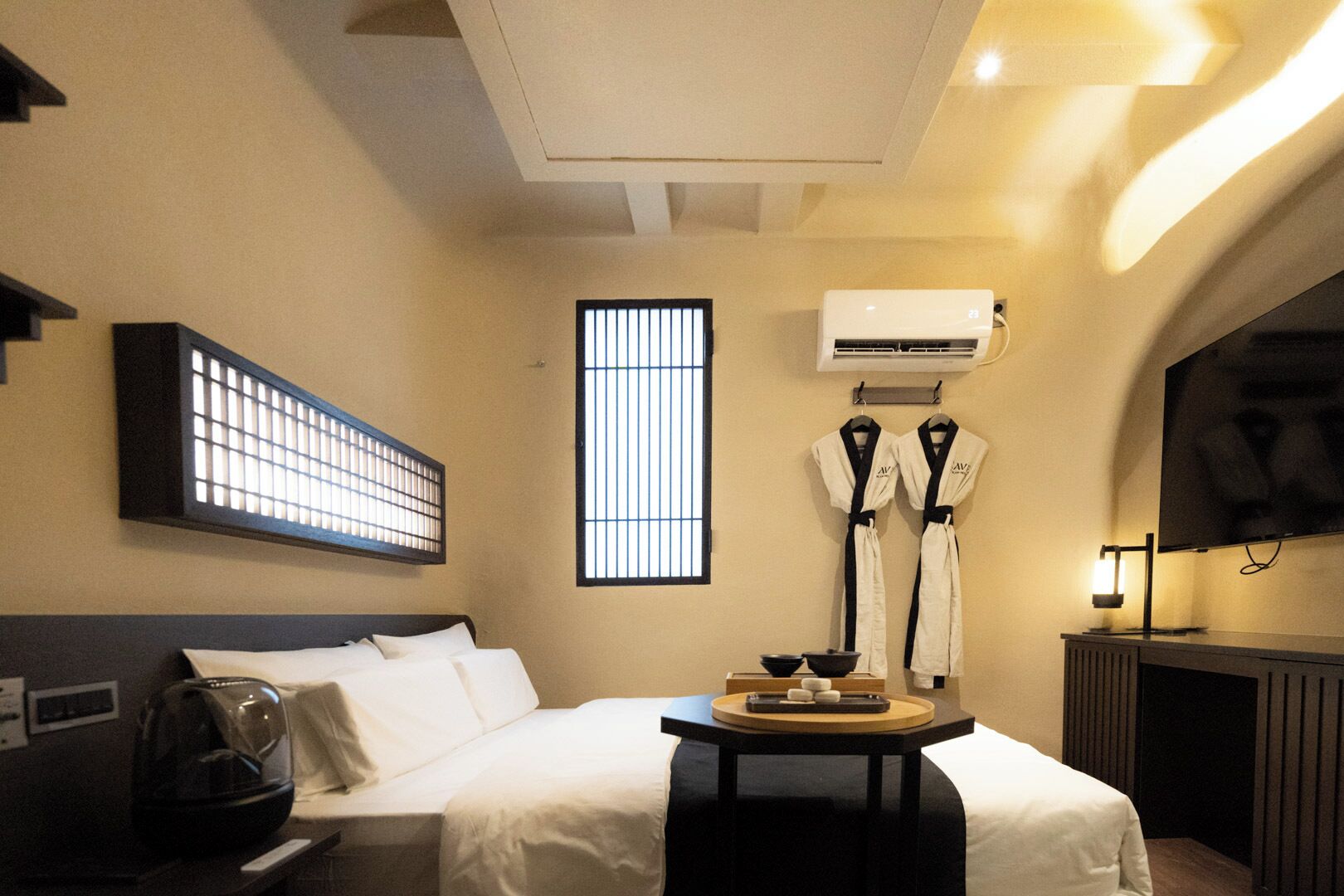 A hotel room with a bed, a massage table, a window with bars, an air conditioning unit, and a television.