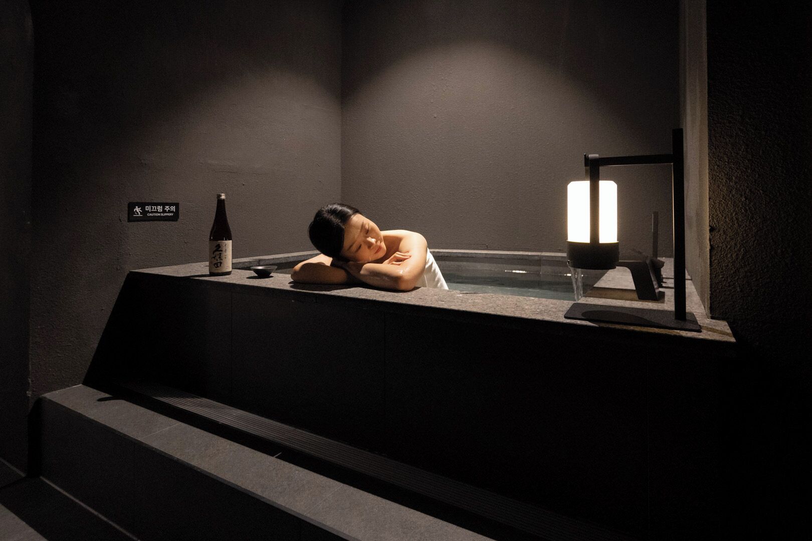 A person relaxing in a dimly lit room with a caution sign, a bottle, and a small table.