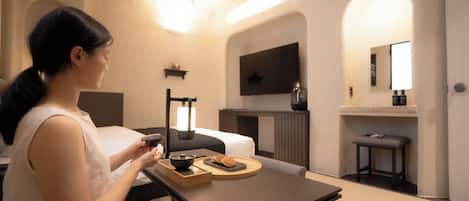 A modern hotel room with a bed, a bedside table with a lamp, a flat-screen TV mounted on the wall, and a small seating area with a table and chairs.