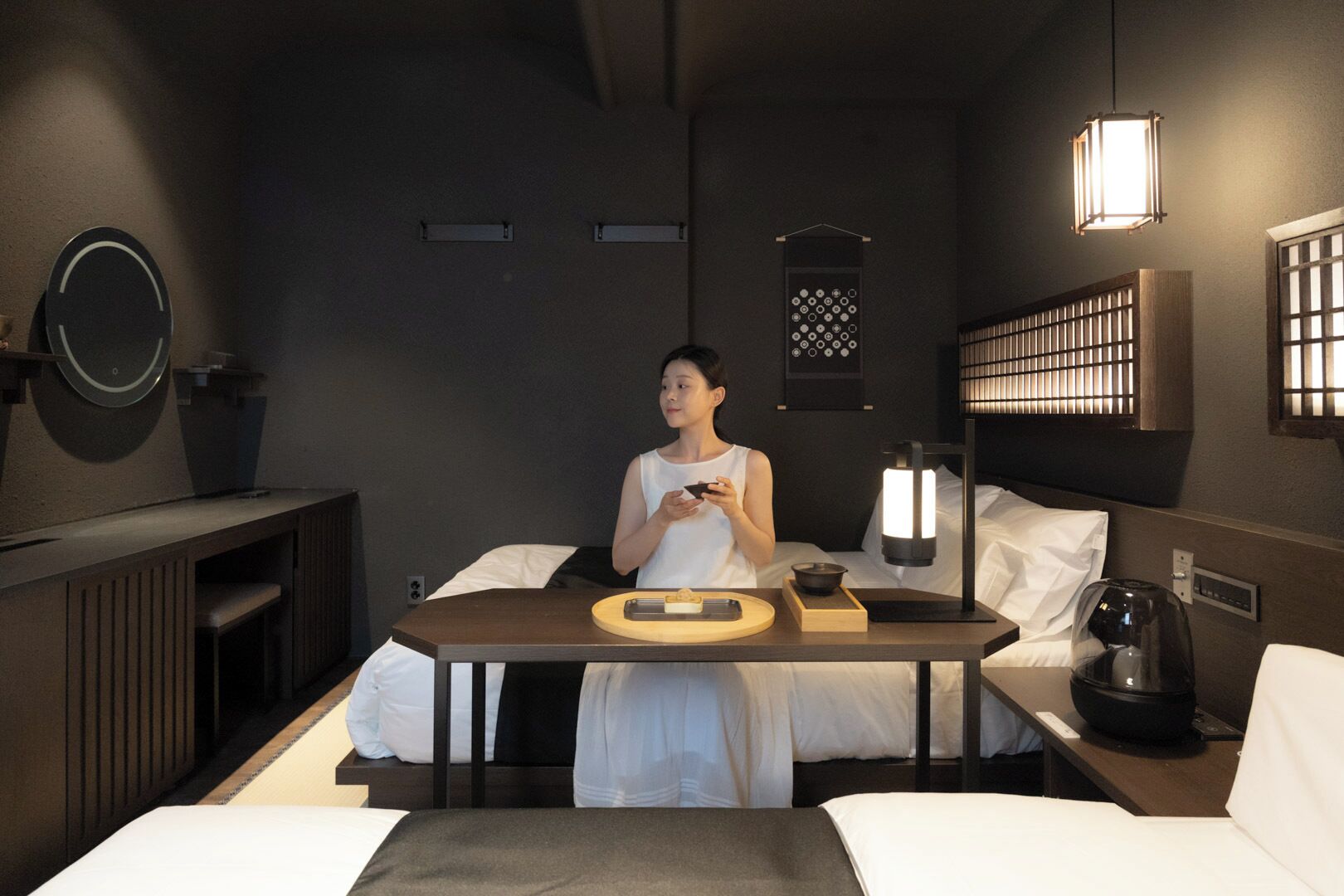 A modern hotel room with a bed, bedside table, lamp, and a small table with a bowl and a remote control.