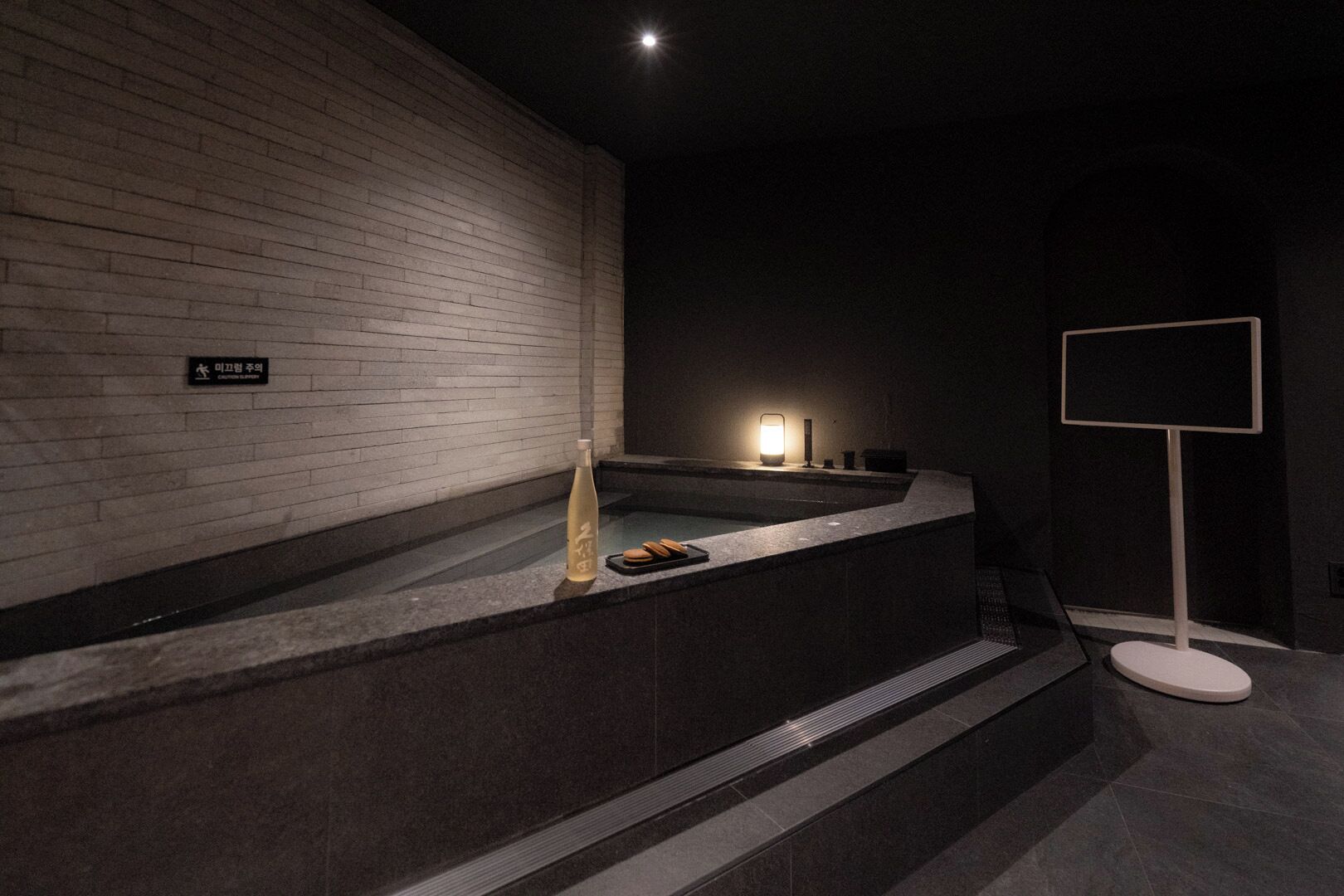 Ein modernes Badezimmer mit Backsteinwand, einer Badewanne, einem kleinen Tisch mit Flasche und Keksen sowie einem Schild in einer Fremdsprache.