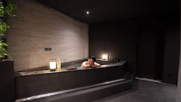 Zona de spa con una persona descansando en una bañera, una luz empotrada en la pared y una botella sobre la encimera.