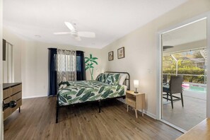 A bedroom with a bed, a ceiling fan, a nightstand with a lamp, a window with curtains, and a view of an outdoor pool area. - Heated Pool No Extra Fee ! firepit| Game Room (Palm Coast)