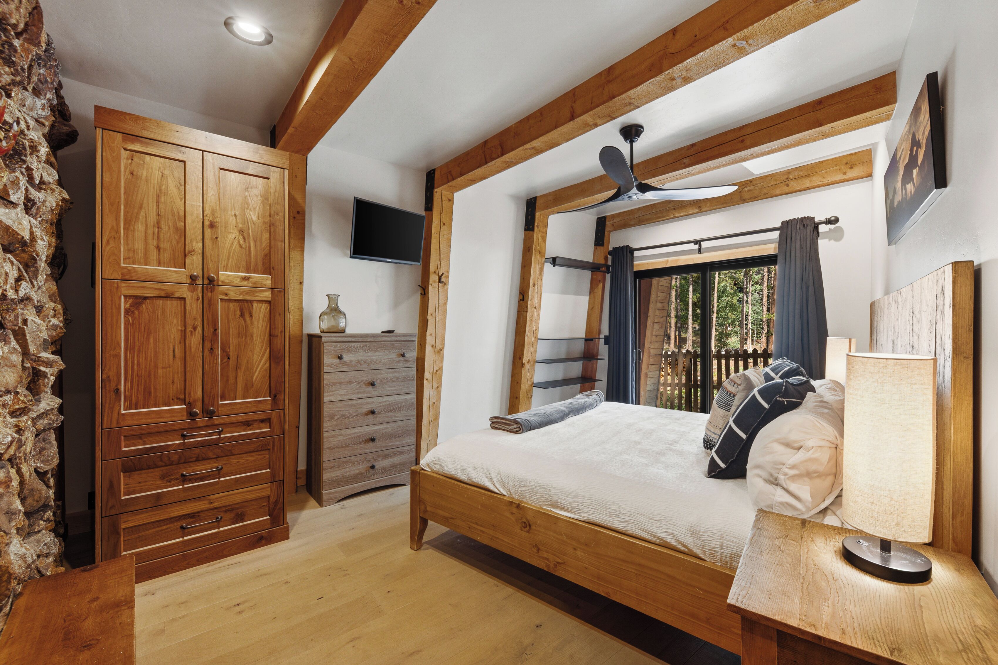 A bedroom with a wooden bed, a dresser, a television, and a window with a view of trees.