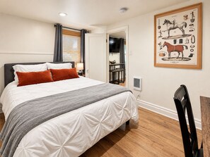 Ein Schlafzimmer mit einem Bett, einem Nachttisch, einer Lampe, einem gerahmten Bild an der Wand und einer Tür zu einem anderen Raum.