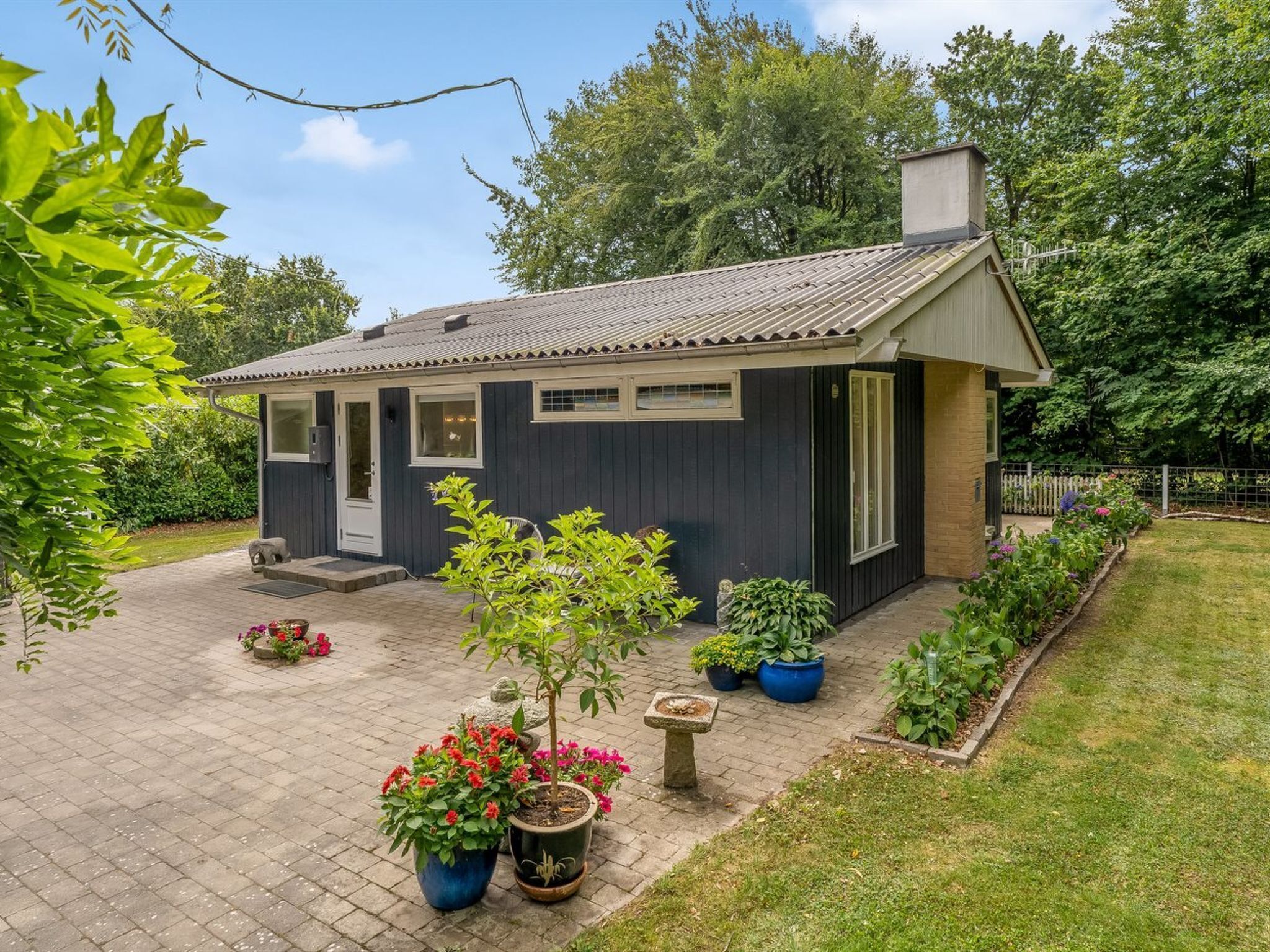 A small, dark-colored house with a tiled roof, a front door, and a chimney. There are potted plants and a small garden area with a pathway leading to the house.