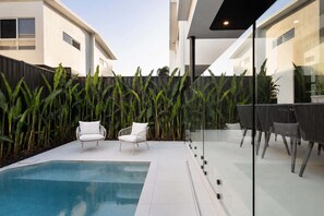 A modern outdoor pool area with a glass partition, two white chairs, and a dining table with chairs.