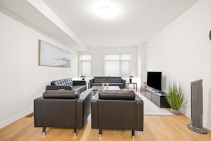 A modern living room with a sectional sofa, a flat-screen TV, and a large window with blinds. - PINE: Private Room with Private Bathroom in Toronto, Canada (Toronto)