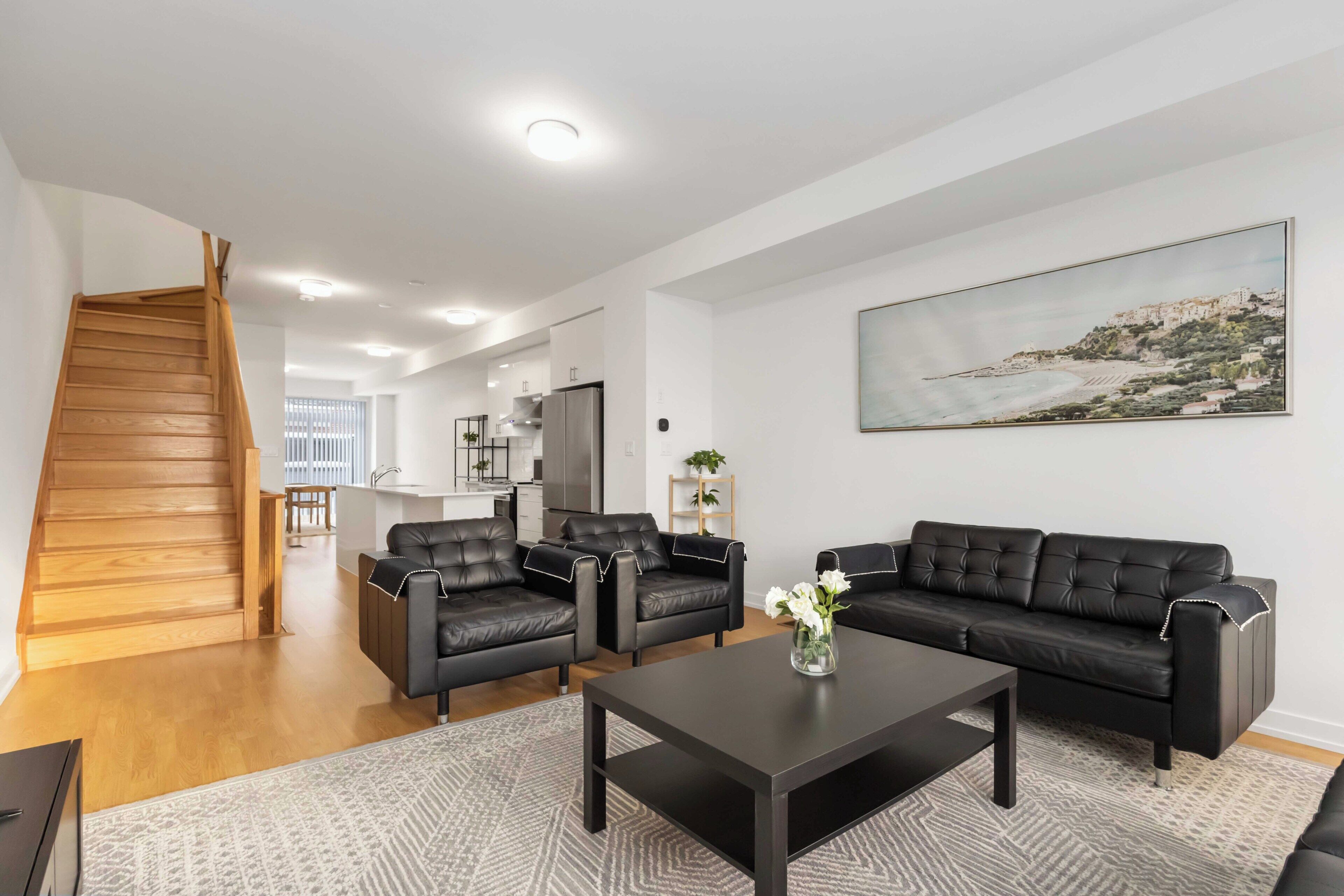 A modern living room with a black leather sofa set, a coffee table, and a painting of a coastal landscape.