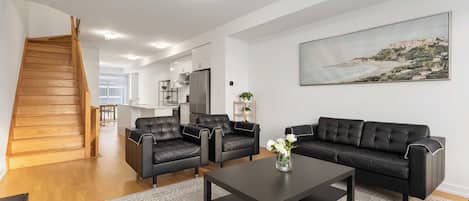 A modern living room with a black leather sofa set, a coffee table, and a painting of a coastal landscape.