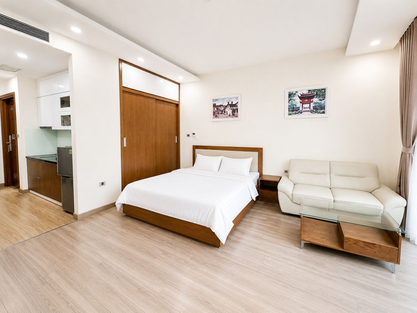 A modern hotel room with a bed, a sofa, a glass coffee table, and a kitchenette.