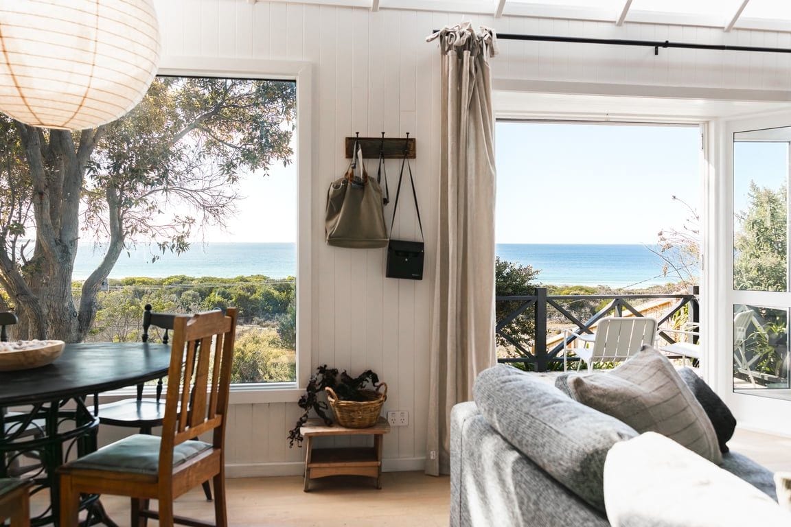 A room with a dining table, chairs, a sofa, a view of the sea, and a hanging bag.