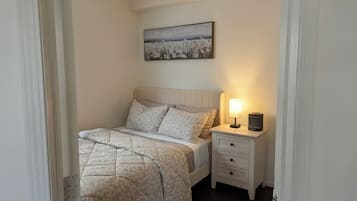 Ett sovrum med en säng, ett sängbord med en lampa, en låda och en inramad tavla på väggen.