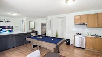 Ett vardagsrum med ett biljardbord, en soffa, en tv och en köksdel med träskåp och en diskmaskin.