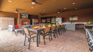 An outdoor dining area with brick flooring, multiple tables and chairs, a stone fireplace, and ceiling fans. - "The Ultimate" 1.3 Acre! Guesthouse & Massive Pool! Pickleball & Putting Greens! (Gilbert)