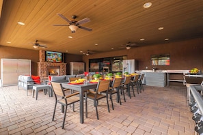 An outdoor dining area with brick flooring, multiple tables and chairs, a stone fireplace, and ceiling fans. - "The Ultimate" 1.3 Acre! Guesthouse & Massive Pool! Pickleball & Putting Greens! (Gilbert)