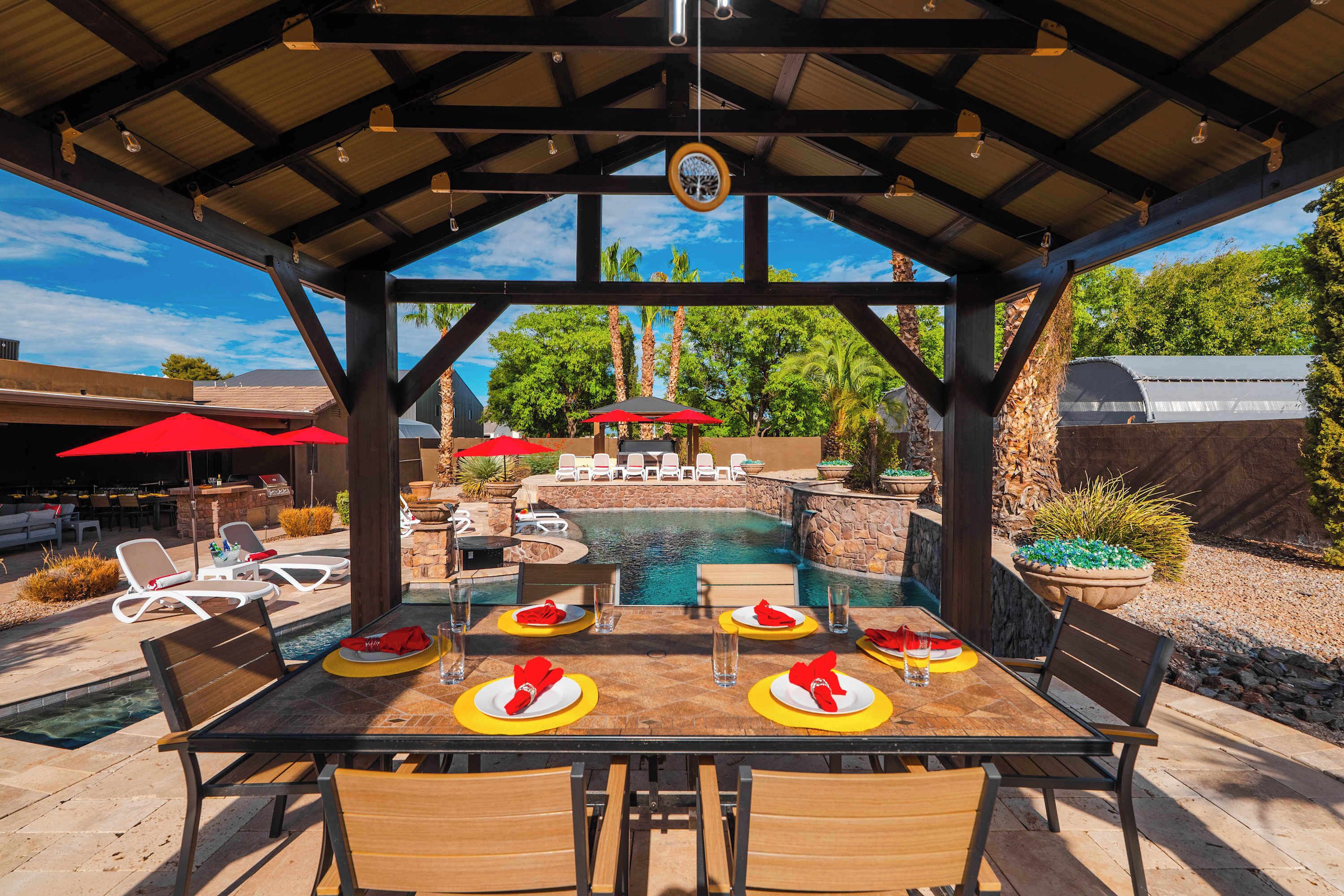 An outdoor pool area with a covered seating area, lounge chairs, and a dining table set for four.