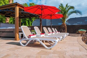 A row of white lounge chairs with red cushions, a red umbrella, and a wooden pergola structure. - "The Ultimate Escape" over 1 Acre w/ Massive Pool!  Pickleball, & Putting Green! (Gilbert)