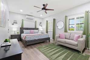 A modern bedroom with a bed, bedside tables, a ceiling fan, an air conditioning unit, a sofa, a mirror, and a window with blinds. - "The Ultimate Escape" over 1 Acre w/ Massive Pool!  Pickleball, & Putting Green! (Gilbert)