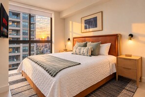 Ein Hotelzimmer mit einem großen Bett, Blick auf die Stadt, einem gerahmten Bild an der Wand und einem Nachttisch mit Lampe.