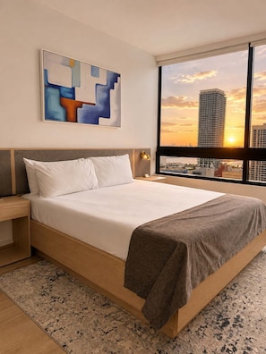 Hotelkamer met een groot bed, nachtkastje, een abstract schilderij aan de muur en uitzicht op een stadshemel bij zonsondergang.