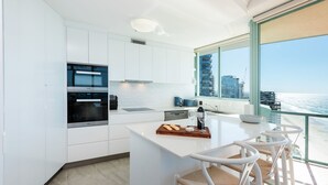 A modern kitchen with white cabinetry, a built-in oven, and a large window overlooking the ocean. - 3 Bedroom Skyview Sub Penthouse (Main Beach)