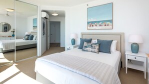 A bedroom with a large bed, two bedside tables with lamps, a ceiling fan, a large mirror, and a beach-themed painting on the wall. - 3 Bedroom Skyview Sub Penthouse (Main Beach)