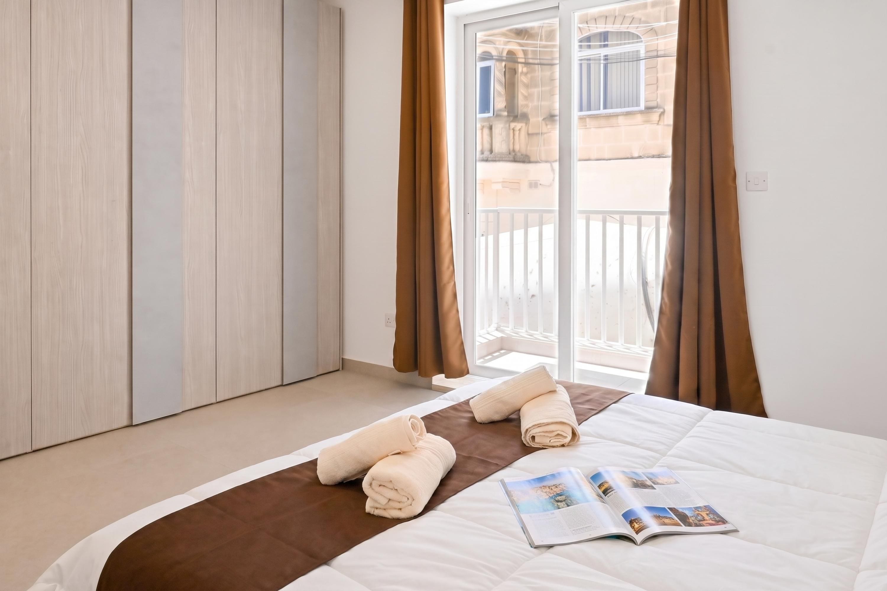 A hotel room with a bed, a magazine, and a view of the outside.