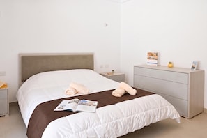 A neatly made bed with a white comforter and a brown blanket, a small bedside table with a book and a plant, and a grey dresser with a magazine on top. - Pool And Sea - Modern Apart Sannat Gozo (Sannat)
